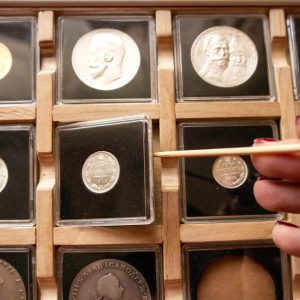 Female hand taking out a coin from a wooden display case with numismatic collection with a wooden stick. Coin holder case with plastic protection square capsules